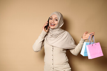 Attractive Muslim Arab woman with beautiful smile in hijab holding color paper bags and talking on mobile phone.