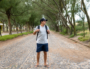 Brunette man on vacation traveling carrying a backpack on his back wearing slippers and a cap on a street beside the sea with cobblestones and large trees that form a green tunnel.