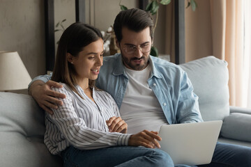 Wall Mural - Close up man and woman using laptop, relaxing on couch together, looking at screen, browsing apps, surfing internet, making video call to friends or relatives, spending leisure time with computer