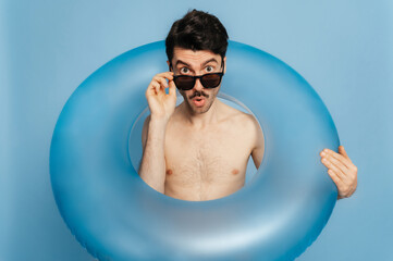 Amazed excited shirtless cheerful caucasian foolish young man in a sunglasses looks surprised through inflatable ring at the camera, stands over blue isolated background, summer, beach time