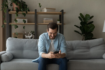 Wall Mural - Smiling man wearing glasses having fun with phone at home, sitting on couch, looking at mobile device screen, chatting or shopping online, browsing apps, enjoying leisure time with smartphone