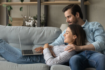 Wall Mural - Side view close up smiling woman using laptop, lying on boyfriend laps, relaxing on couch at home, happy young couple looking at computer screen, shopping or chatting online, enjoying leisure time