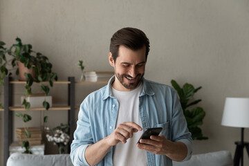 Close up smiling man using phone, browsing apps, having fun, standing at home, holding smartphone, happy satisfied young male wearing glasses chatting or shopping online, enjoying leisure time