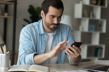 Wall Mural - Close up confident man wearing glasses looking at smartphone screen, sitting at work desk with laptop, browsing apps, writing message in social network, customer shopping or chatting online