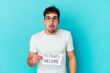 Wall Mural - Young caucasian man holding a Refugees welcome placard isolated shrugs shoulders and open eyes confused.