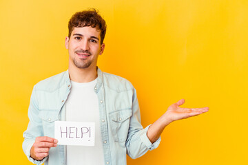 Wall Mural - Young caucasian man holding a help placard isolated showing a copy space on a palm and holding another hand on waist.