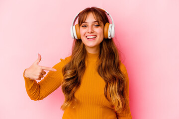 Wall Mural - Young caucasian woman listening music with headphones isolated person pointing by hand to a shirt copy space, proud and confident