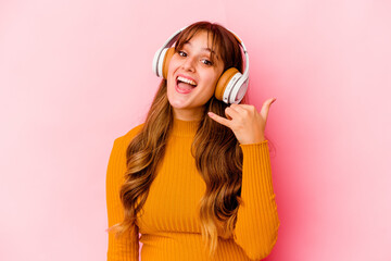 Wall Mural - Young caucasian woman listening music with headphones isolated showing a mobile phone call gesture with fingers.