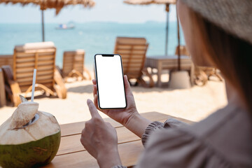 Mockup image of a woman holding mobile phone with blank desktop screen while sitting on the beach