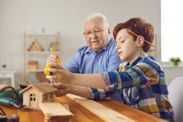 Grandfather and teen grandson making wooden toy houses. Old carpenter teaching little boy new handwork skills. Senior man and child with pencils behind ears working with wood in carpentry workshop