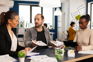 Wall Mural - Worried startup diverse team discussing about financial results, discussing sitting at desk in office, planning next strategy holding tablet searching solutions. Multiethnic businesspeople working