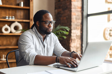 Busy bored young African-American male author office worker in glasses sitting at the desk typing on laptop, working on project, writing creating article or blog, feeling lack of motivation to finish