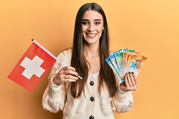 Wall Mural - Beautiful brunette young woman holding switzerland flag and franc banknotes smiling with a happy and cool smile on face. showing teeth.