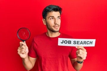 Young handsome man holding magnifying glass for job search smiling looking to the side and staring away thinking.