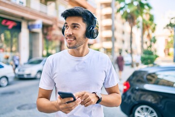 Wall Mural - Young latin man smiling happy using smartphone and headphones at the city.