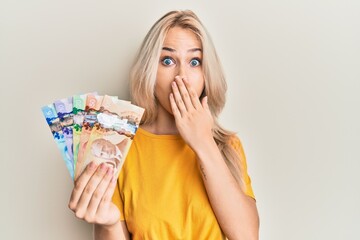 Canvas Print - Beautiful caucasian blonde girl holding canadian dollars covering mouth with hand, shocked and afraid for mistake. surprised expression