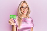 Fototapeta  - Beautiful caucasian blonde girl holding birth control pills looking positive and happy standing and smiling with a confident smile showing teeth