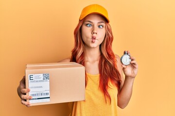 Poster - Young redhead woman holding delivery package and stopwatch making fish face with mouth and squinting eyes, crazy and comical.