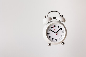 Small white alarm clock, black numbers, set the time for 10.10 o'clock, placed on a white table.