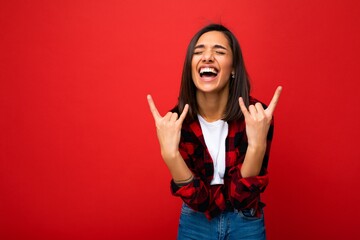 Photo of young happy positive cool attractive brunette woman with sincere emotions wearing white t-shirt and hipster red check shirt isolated on red background with empty space and showing rock sign