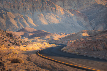 Wall Mural - Dramatic golden light on an empty winding desert road through the rugged terrain of the badlands landscape in Death Valley Park National Park, USA.