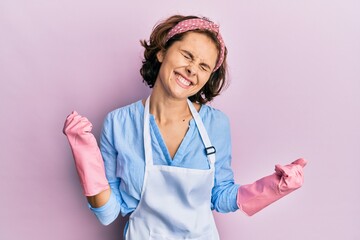 Sticker - Young brunette woman wearing cleaner apron and gloves very happy and excited doing winner gesture with arms raised, smiling and screaming for success. celebration concept.