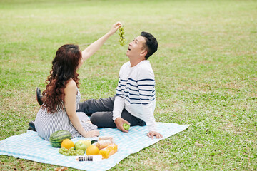 Wall Mural - Pretty young woman feeding boyfriend with grapes when they are sitting on blanket in city park