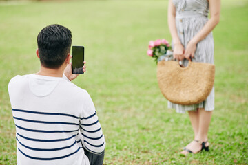 Poster - Young man taking photo of his girlfriend with wicker bag of pink roses in city park