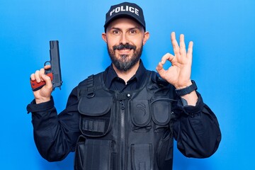 Poster - Young handsome man wearing police uniform holding gun doing ok sign with fingers, smiling friendly gesturing excellent symbol