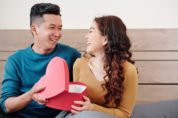 Wall Mural - Happy young woman opening present in heart shape box from her boyfriend when sitting on bed in the morning