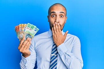 Canvas Print - Hispanic adult man holding australian dollars covering mouth with hand, shocked and afraid for mistake. surprised expression