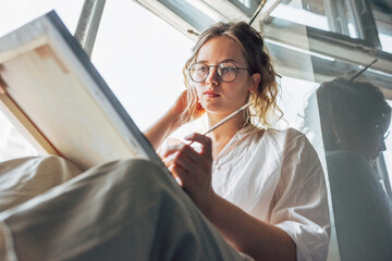 Wall Mural - Female artist painting on canvas in her art studio sitting next to the window. A woman painter with eyeglasses painting with oil searching for imagination in the workshop.