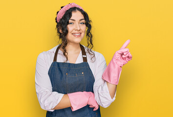 Wall Mural - Young hispanic girl wearing cleaner apron and gloves smiling happy pointing with hand and finger to the side