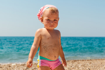 beautiful baby girl of one year old in pink bathing trunks stands on the beach in the summer. sand is glued to the child's body