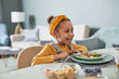 © Seventyfour - Portrait of cute African-American girl sitting at dinner table and laughing while enjoying meal in cozy home interior, copy space