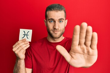 Poster - Young caucasian man holding paper with pisces zodiac sign with open hand doing stop sign with serious and confident expression, defense gesture