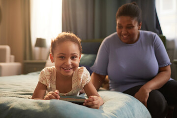 Portrait of cute African-American girl laying on bed and smiling at camera happily with mother in background, copy space