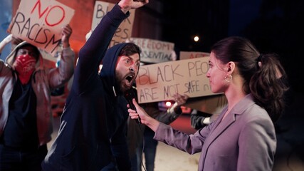 Wall Mural - Caucasian female correspondent interviewing aggressive male protester at street at night while people protesting for human rights and against police violence. Interview of rebel man by woman for news.