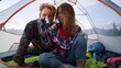 Young couple sitting in tent during hike. Redhead guy drinking tea from cup