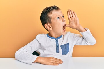 Wall Mural - Adorable caucasian kid wearing casual clothes sitting on the table shouting and screaming loud to side with hand on mouth. communication concept.