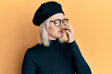 Poster - Young blonde girl wearing french look with beret smelling something stinky and disgusting, intolerable smell, holding breath with fingers on nose. bad smell