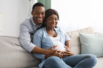 Wall Mural - Smiling black man hugging his pregnant woman sitting on sofa