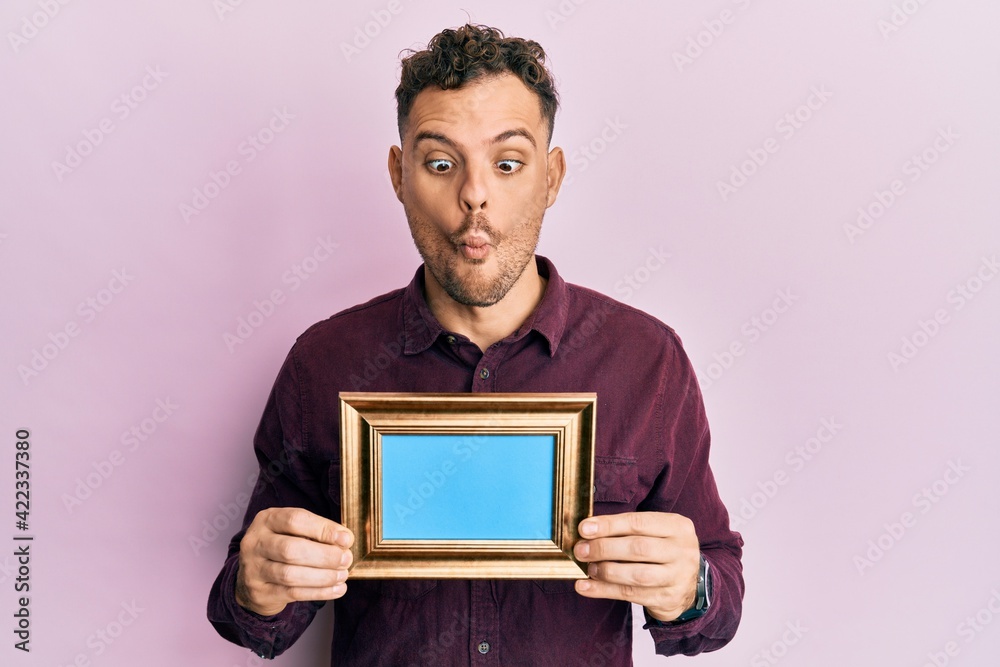 Young hispanic man holding empty frame making fish face with mouth and squinting eyes, crazy and comical.
