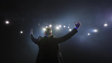 a man in a business suit is going on the stage. spotlights are shining on him. he is holding his han