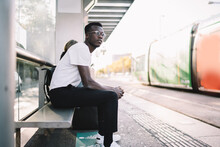Serious Dark Skinned Male In Eyewear Sitting On Bus Stop In Shelter Waiting For Publica Transport, Handsome Casually Dressed Hipster Guy Looking Away For Tramway Spending Time On Urban Settings
