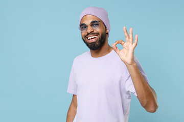 Wall Mural - Young smiling happy positive satisfied hipster fun unshaven black dark-skinned african man 20s wear violet t-shirt hat glasses show ok okay gesture isolated on pastel blue background studio portrait.