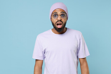 Wall Mural - Young surprised shocked astonished impressed unshaven student black african man 20s in violet t-shirt hat glasses looking camera with opened mouth isolated on pastel blue background studio portrait.