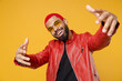 Young african cool rapper happy man 20s with funky trendy pink hairdo wearing stylish red leather jacket glasses Stretching hands to camera isolated on yellow orange color background studio portrait.