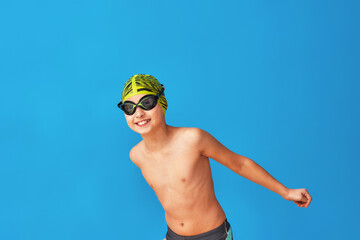 Wall Mural - portrait of a sporty boy in a swimming cap, glasses and a bathing suit, performing exercises on a blue background. child enjoys swimming in a sports school. concept healthy lifestyle and sports.