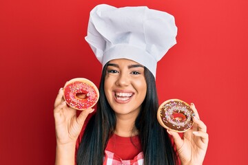 Wall Mural - Young beautiful hispanic girl wearing chef hat holding homemade doughnut smiling and laughing hard out loud because funny crazy joke.